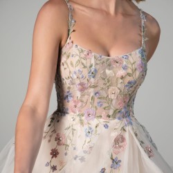 Woman in a sleeveless dress with pastel floral embroidery and sheer tulle overlay, holding the skirt—perfect for those seeking Spring Wedding Dresses or elegant Spring Bridal Gowns.