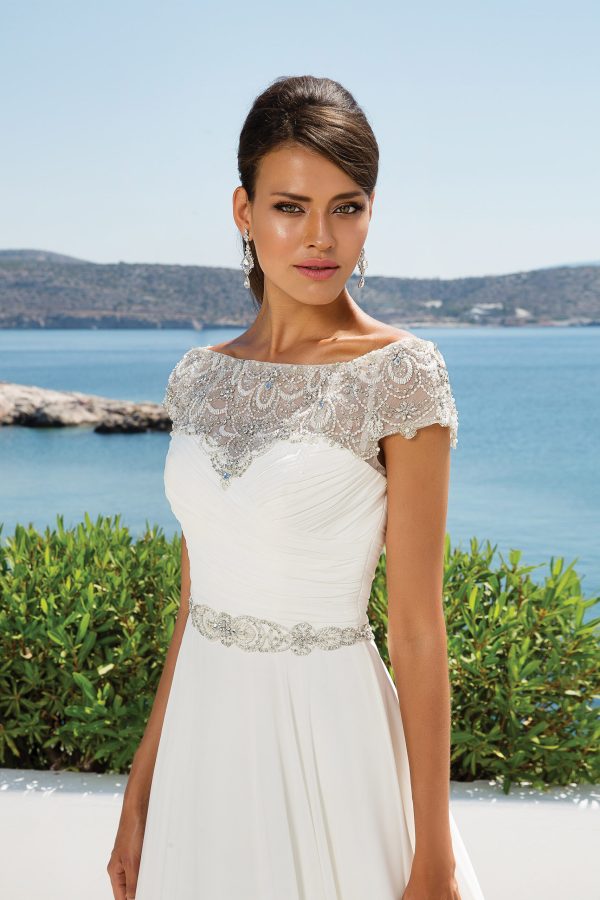 A bride in a white gown with intricate lace detailing on the bodice and cap sleeves stands in front of a scenic coastal backdrop with blue water and distant hills. With the poise of a Greek Goddess, she wears long earrings and a belt with embellishments at the waist, looking directly at the camera.