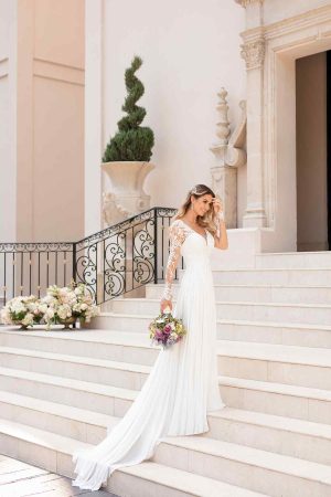A happy bride in a pretty, white, long-sleeved lace wedding dress descends the steps of a grand building holding a bouquet of flowers. The building has ornate details, a large potted plant, and a decorative wrought iron railing. She appears to be touching her hair lovingly.