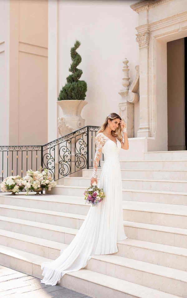 A happy bride in a pretty, white, long-sleeved lace wedding dress descends the steps of a grand building holding a bouquet of flowers. The building has ornate details, a large potted plant, and a decorative wrought iron railing. She appears to be touching her hair lovingly.