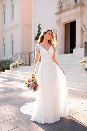 A happy woman in a long white bridal gown with lace sleeves holds a bouquet of flowers. She stands outdoors on a pathway with a staircase and an ornate building in the background, smiling and looking towards the right. Sunlight illuminates her and the pretty surroundings, making the moment filled with love.