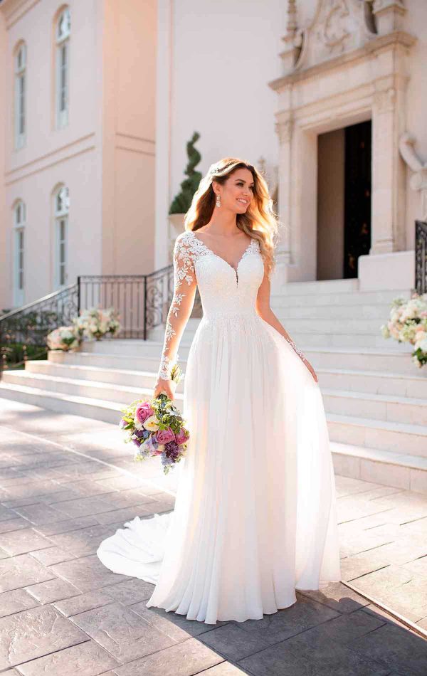 A happy woman in a long white bridal gown with lace sleeves holds a bouquet of flowers. She stands outdoors on a pathway with a staircase and an ornate building in the background, smiling and looking towards the right. Sunlight illuminates her and the pretty surroundings, making the moment filled with love.