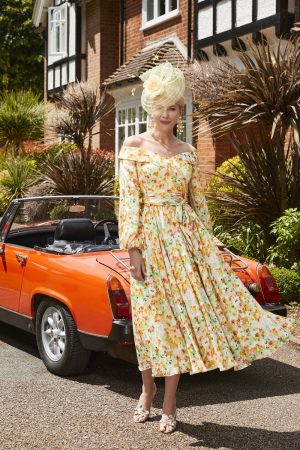 A woman in a Veni Infantino 29664A floral off-shoulder dress and yellow hat stands next to an orange convertible car parked in front of a house with Tudor-style architecture. She is smiling, and the weather appears sunny and clear.