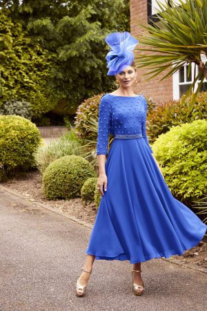 A woman stands outdoors on a paved path, wearing a striking royal blue Veni Infantino - 29825 dress adorned with pearl embellishments on the bodice and sleeves. She completes the look with gold heels and a large blue fascinator. The background is lush with greenery and manicured bushes.