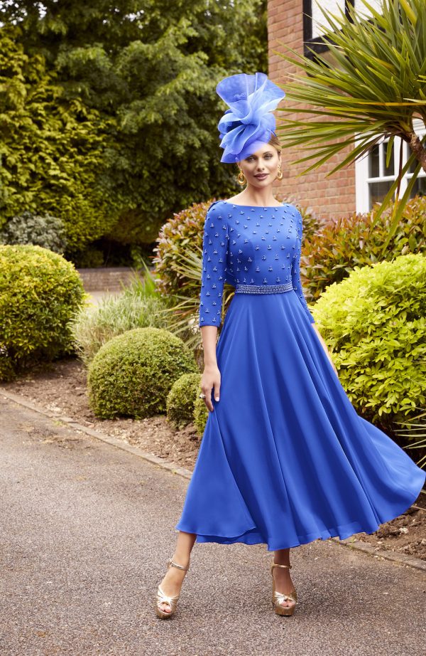 A woman stands outdoors on a paved path, wearing a striking royal blue Veni Infantino - 29825 dress adorned with pearl embellishments on the bodice and sleeves. She completes the look with gold heels and a large blue fascinator. The background is lush with greenery and manicured bushes.