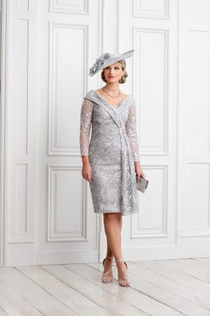 A woman stands in front of a white paneled wall, wearing the stunning Ispirato - Slate dress that features intricate lace details and falls to knee-length. Completing her elegant look is a matching wide-brimmed hat adorned with a flower and ISH814 silver high-heeled sandals. She accessorizes with a small slate-colored clutch and stylish jewelry.