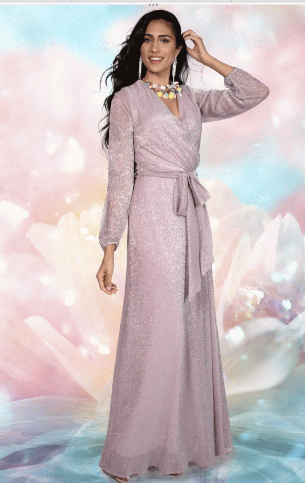 A woman in a shimmering, long-sleeve Frank Lyman - 228212 pale pink gown with a sash at the waist, posing with one hand on her head. The background is ethereal and colorful, resembling a soft-focus floral and light motif. Feeling like she's part of painting number 228212—simply enchanting.