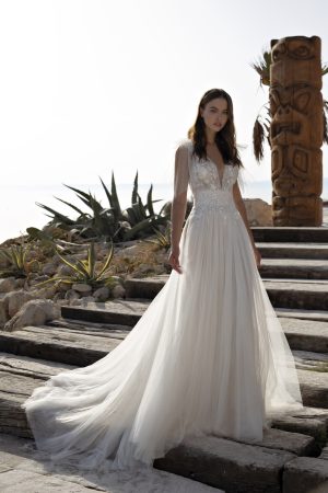 A woman in a flowing, white bridal gown from the "Papillon by Modeca" collection, specifically the Ohanna dress, with intricate lace details stands on outdoor wooden steps. The dress features a deep V-neck and sheer sleeves. Behind her are plants, rocks, and a wooden totem pole against a bright, hazy sky.