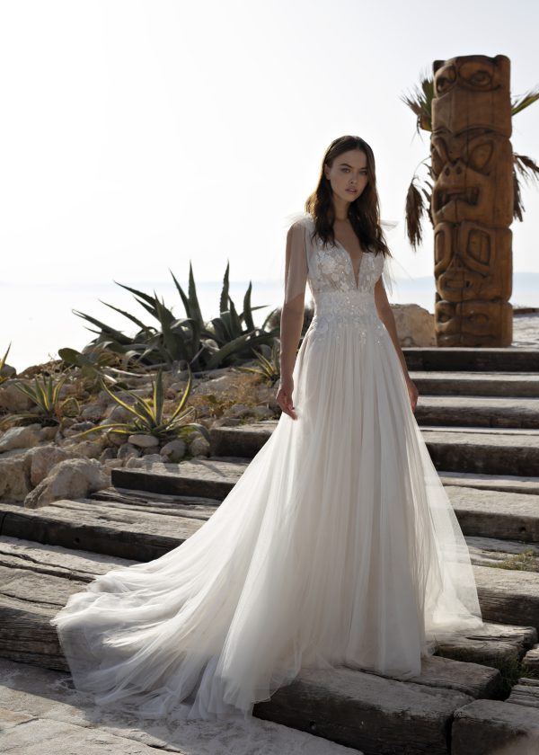 A woman in a flowing, white bridal gown from the "Papillon by Modeca" collection, specifically the Ohanna dress, with intricate lace details stands on outdoor wooden steps. The dress features a deep V-neck and sheer sleeves. Behind her are plants, rocks, and a wooden totem pole against a bright, hazy sky.