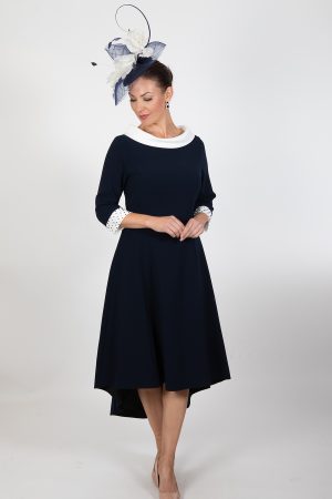 A woman wearing the Lizabella - Ella Boo - 2403 dress in navy blue with a white collar and cuffs stands against a plain white background. She accessorizes with a navy blue fascinator adorned with white and navy feathers, beige heels, and turquoise earrings, her hands clasped in front of her.