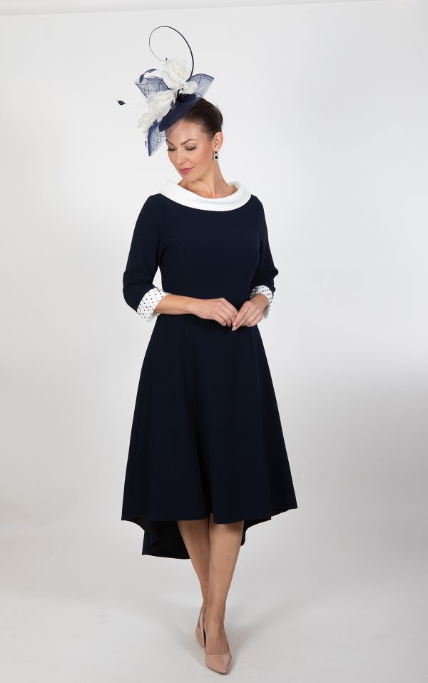 A woman wearing the Lizabella - Ella Boo - 2403 dress in navy blue with a white collar and cuffs stands against a plain white background. She accessorizes with a navy blue fascinator adorned with white and navy feathers, beige heels, and turquoise earrings, her hands clasped in front of her.