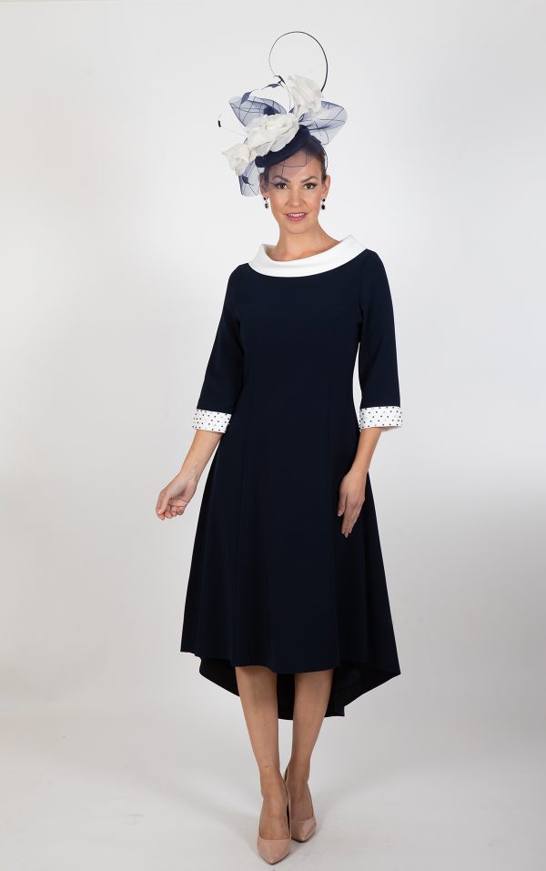 A woman is wearing the Lizabella - Ella Boo - 2403 dark blue dress, which features a high-low hemline, white collar, and lace cuffs. She complements her attire with a large decorative blue and white hat and finishes her look with nude heels. Smiling, she stands in front of a plain background.