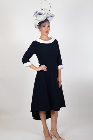 A woman stands against a plain white background, adorned in an elegant Lizabella - Ella Boo - 2403 navy blue dress with white accents and a high-low hemline. She accessorizes with a stylish large fascinator hat embellished with white and navy blue elements, completing her ensemble with beige heels.