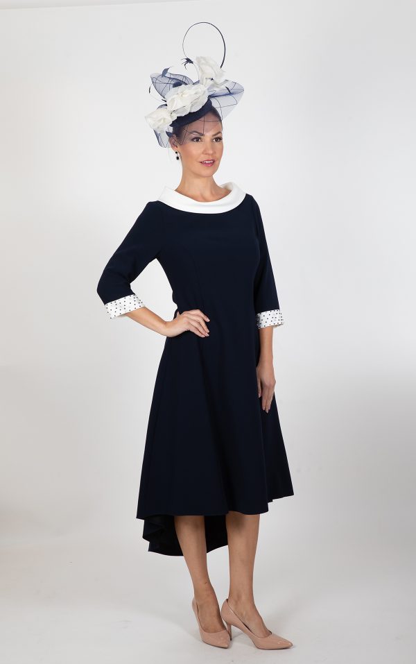 A woman stands against a plain white background, adorned in an elegant Lizabella - Ella Boo - 2403 navy blue dress with white accents and a high-low hemline. She accessorizes with a stylish large fascinator hat embellished with white and navy blue elements, completing her ensemble with beige heels.