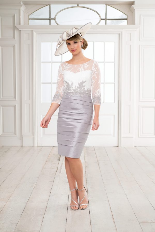 A woman stands in a bright room with white-paneled walls and a large arched window. She wears a stylish, knee-length silver Ispirato dress with a lace top and sheer sleeves, accessorized by an elegant large hat. Her sophisticated look is completed with high-heeled ISH909 sandals from Ispirato.