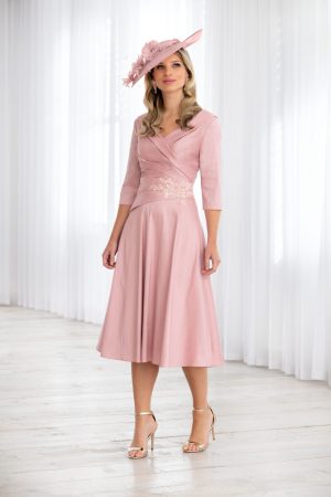 A woman stands in an elegantly draped room, wearing a light pink, knee-length dress with three-quarter sleeves and floral embroidery at the waist. She accessorizes with the matching Ispirato - ISH822 pink hat adorned with flowers and wears gold high-heeled sandals.