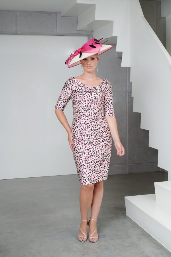 A woman stands indoors wearing a fitted, knee-length Ispirato - ISJ828 dress with a pink, black, and white polka-dot pattern. She pairs the dress with a large pink hat accented with black and pink feathers and is wearing strappy high-heeled shoes. The background features a modern, minimalistic staircase.