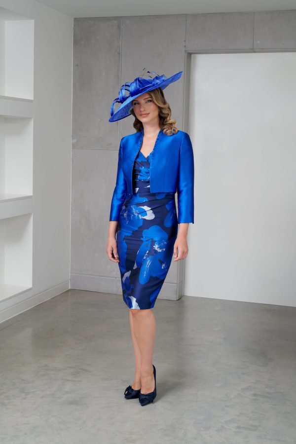 A woman stands indoors wearing a stylish blue outfit from Ispirato - ISJ900. She has on a blue floral dress paired with a matching cropped jacket and an elaborate hat adorned with feathers. Wearing dark shoes, she poses against the minimalistic backdrop of white walls and shelves.