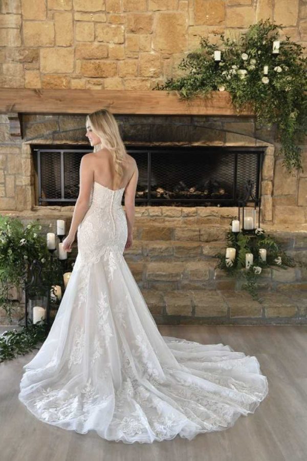 A bride in a strapless Stella York - 7397 - Peony lace wedding dress stands with her back to the viewer, facing a stone fireplace adorned with greenery and white candles. The dress has a long, flowing train that spreads out behind her on the wooden floor.