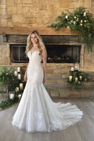 A woman in a strapless, elegant white Stella York 7397 - Peony wedding gown with a fitted bodice and flared train stands in front of a stone fireplace adorned with peony greenery and candles. She has long, wavy hair and is smiling slightly. The setting has a rustic, cozy ambiance.