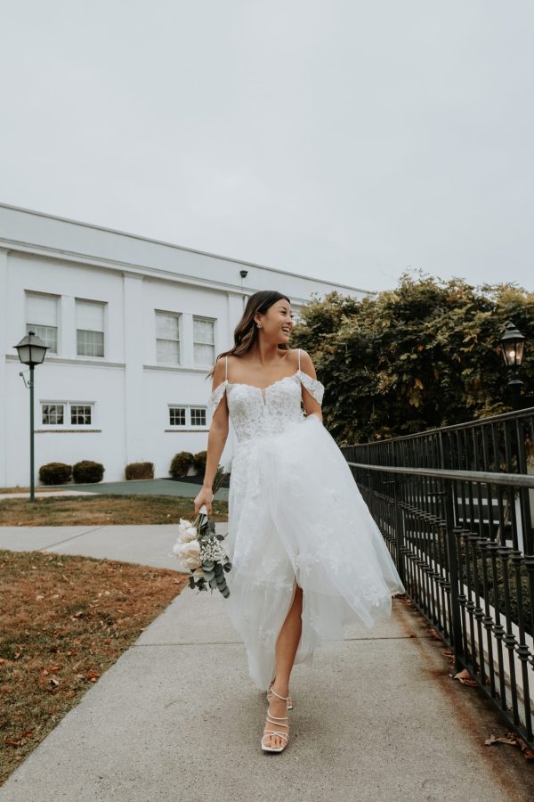 A bride in a white Stella York Bali 7447 wedding dress walks outside near a building, holding a bouquet of flowers and smiling. She has long, dark hair and is wearing strappy heels. The dress features a lace bodice and flowing skirt. Bushes and an old-fashioned lamp post are in the background.