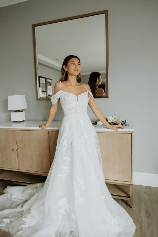 A bride stands confidently in a room wearing a stunning Stella York - Bali - 7447 white off-shoulder, lace wedding gown. She has a serene expression, with one hand resting on a beige console table behind her, and a large square mirror reflecting her image. The room, reminiscent of Bali's serene beauty, is softly lit.