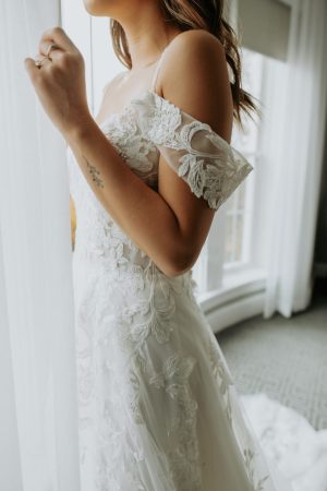 A bride wearing an off-shoulder lace wedding dress stands by a window with sheer curtains. She has one hand raised to touch the curtain, revealing a small tattoo on her inner forearm. Natural light filters through the window, illuminating her Stella York - Bali - 7447 gown, creating an ethereal ambiance.