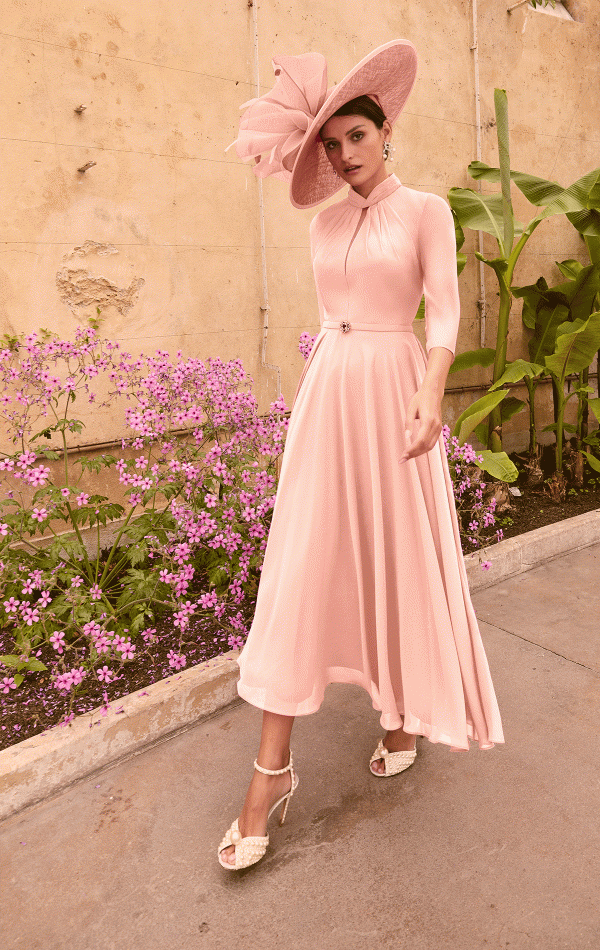 A woman in a flowing pink dress and a large, matching pink hat stands on a pathway. She is wearing strappy high heels and is surrounded by pink flowers and green banana plants, with a beige wall behind her—perhaps she's en route to deliver elegant Invitations by Veni - 36004 for an event.