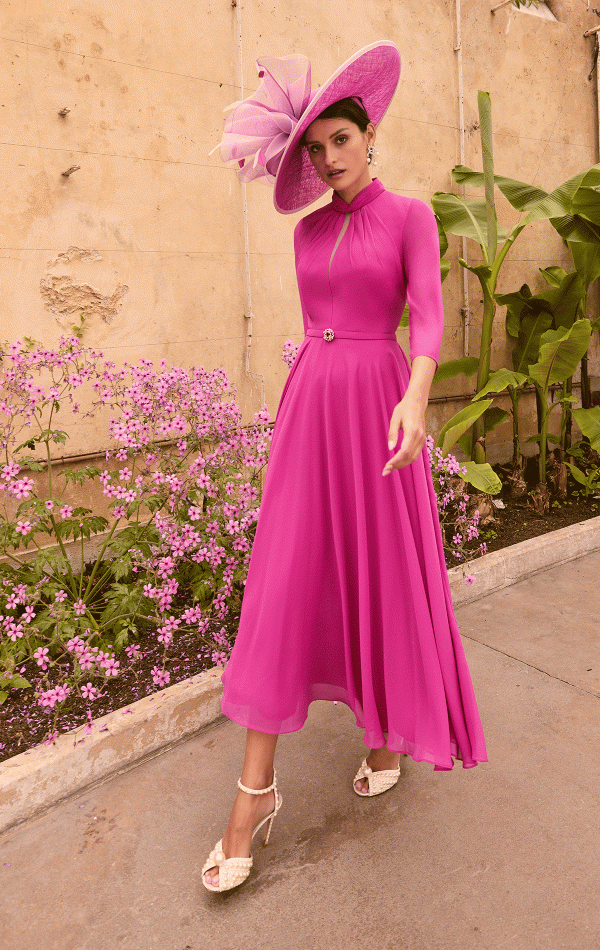 A woman poses in a long-sleeved, fuchsia pink dress by Invitation by Veni - 36004, featuring a flared skirt and cinched waist. She accessorizes with a large matching pink hat adorned with feathers and wears white heels. The background showcases pink flowers, tall plants, and a beige wall.