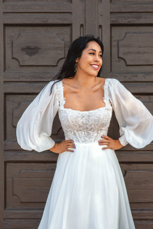 A woman in a white Stella York 7813 - Nala wedding dress with long, sheer, puffed sleeves and intricate lace detailing on the bodice stands in front of a rustic wooden door. She has long dark hair, smiles brightly, and poses with her hands on her hips.