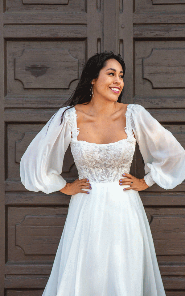 A woman in a white Stella York 7813 - Nala wedding dress with long, sheer, puffed sleeves and intricate lace detailing on the bodice stands in front of a rustic wooden door. She has long dark hair, smiles brightly, and poses with her hands on her hips.