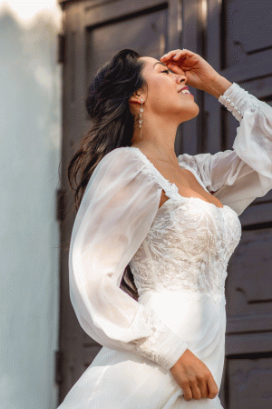 A woman in a white, long-sleeved lace Stella York 7813 - Nala wedding gown is pictured outdoors. She has long, flowing dark hair and is smiling blissfully with her eyes closed, tilting her head slightly back and touching her forehead with one hand. A dark wooden door is visible in the background.