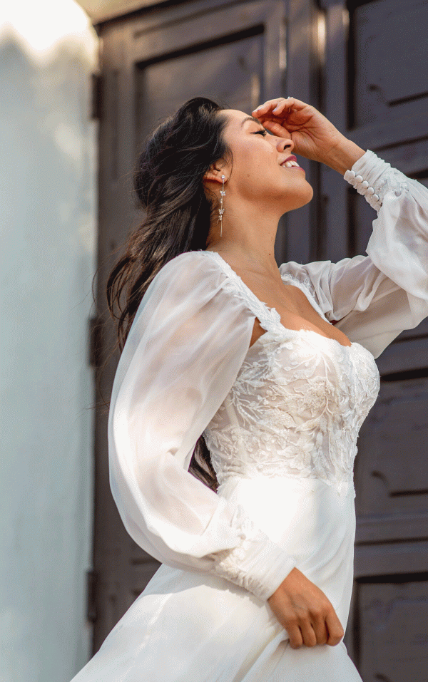 A woman in a white, long-sleeved lace Stella York 7813 - Nala wedding gown is pictured outdoors. She has long, flowing dark hair and is smiling blissfully with her eyes closed, tilting her head slightly back and touching her forehead with one hand. A dark wooden door is visible in the background.