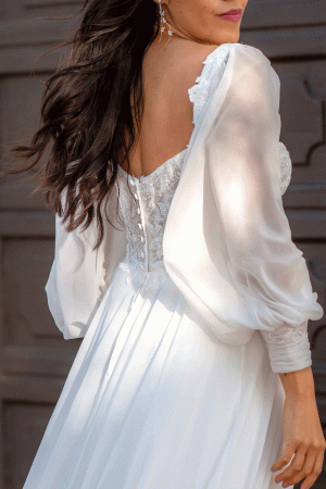 A woman with long, flowing hair is seen from the side wearing an elegant Stella York 7813 - Nala gown, featuring intricate lace detailing on the bodice and sheer, billowy sleeves. She is outdoors against a backdrop of a dark wooden door. This breathtaking dress exudes timeless charm.