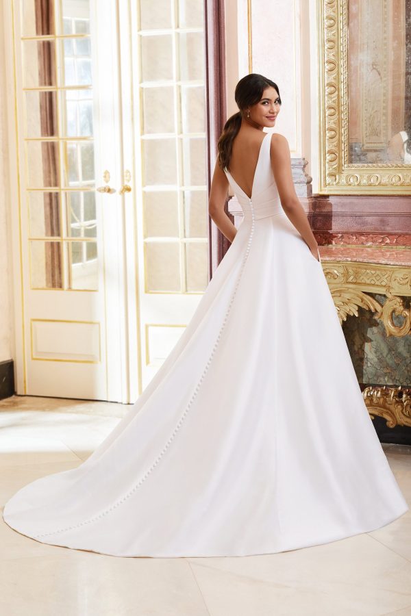 A bride stands in an elegant room, facing away. She is wearing the Justin Alexander 44080 - Dior, a white sleeveless wedding gown featuring a deep V-back and a long, flowing train adorned with buttons. Her hair is styled in a low ponytail as she stands before ornate glass doors and classic decor, reminiscent of timeless elegance.