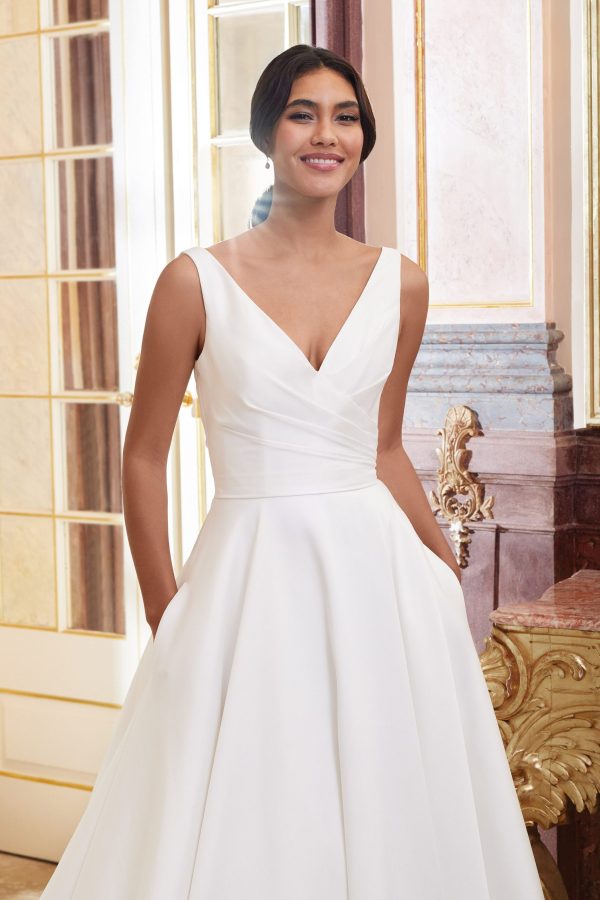 A woman in a white sleeveless wedding dress with a V-shaped neckline, possibly the Justin Alexander 44080 - Dior, poses indoors. The background features elegant decorative elements, including an ornate golden and marble wall and a large window with glass panels. The woman has her hands in her dress pockets.