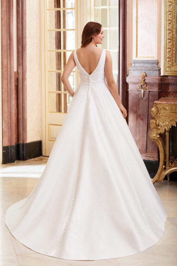 A woman stands with her back to the camera, showcasing a luxurious 44080 - Dior A-line wedding dress. The gown features a deep V-back, a long, flowing train, and a row of buttons running from the lower back to the hem. The setting is an elegant, ornately decorated room.