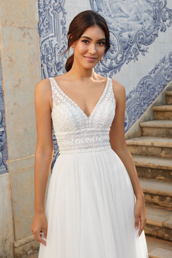 A smiling woman in the stunning Justin Alexander 44120 - Freya wedding dress, featuring intricate lace details, stands in front of a wall adorned with blue and white ornate tiles. She has long, dark hair pulled back, and a set of stone steps are to her right.