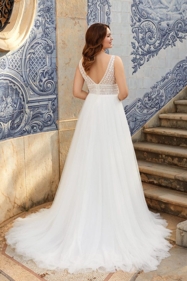 A woman stands with her back to the camera, wearing the Justin Alexander 44120 - Freya, a sleeveless white wedding dress featuring a delicate lace bodice and a flowing tulle skirt. She is in a setting adorned with ornate blue and white tile work on the walls and stone steps to her right.