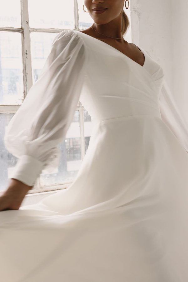 A woman named Eleanore stands in front of a large window, wearing the 44264 - Eleanore wedding dress in white with long, sheer sleeves. The dress features a V-neckline and a flowy skirt. Her face is partially out of frame, and soft light filters through the window behind her.