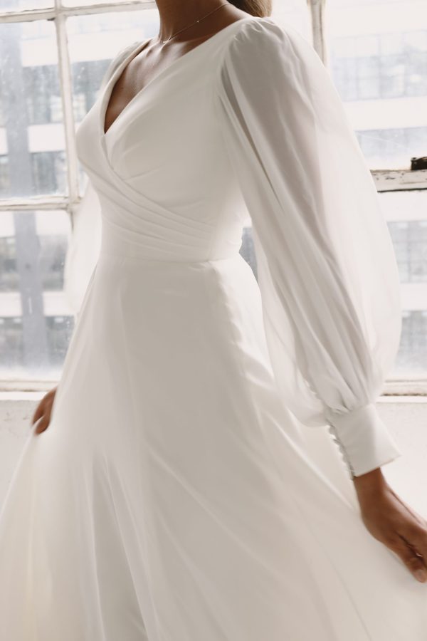 A close-up of Eleanore wearing Product 44264 - the Eleanore wedding dress. The dress features long, sheer, flowing sleeves and a fitted bodice with a V-neckline. The background includes a large window with multiple panes, allowing soft natural light to illuminate the scene.