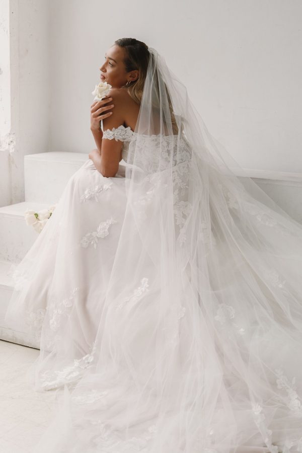 A bride dressed in the Justin Alexander 44303 - Mila off-the-shoulder lace bridal gown with a long lace veil sits on steps holding a white flower. She gazes thoughtfully to the side, with sunlight streaming through a nearby window, creating a serene and elegant scene infused with timeless beauty.