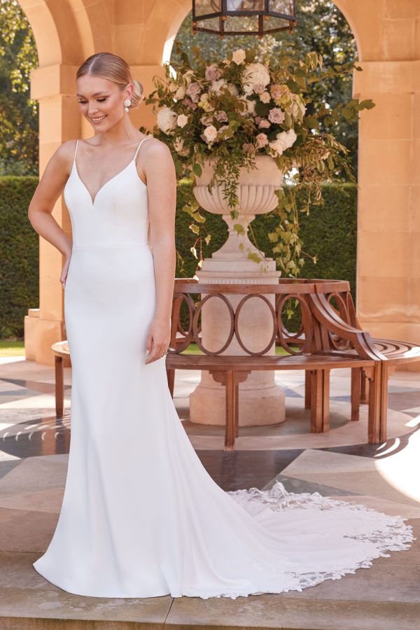 A bride stands outdoors in front of an ornate garden structure, wearing the simple yet elegant Justin Alexander 44322 - Ella gown with a long train. The gown features thin straps and a V-neckline. She smiles while holding one hand on her hip and the other resting by her side. A lush floral arrangement in a stone vase is visible in the background.