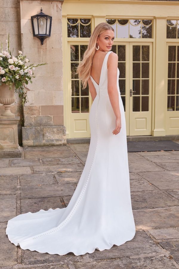 A woman stands on a stone patio in front of a large building with yellow double doors and a lantern. She is dressed in the elegant, backless Justin Alexander 44325 - Anya wedding dress, with a long train, looking to the side. Her blonde hair, styled in loose waves, complements her dangling earrings perfectly.