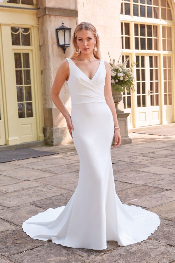 A woman, Anya, stands outside a building with large windows and light yellow doors, wearing the Justin Alexander 44325 - Anya: a white, floor-length, sleeveless gown featuring a v-neckline and fitted bodice. She is accessorized with earrings and a bracelet. The ground is made of stone tiles.