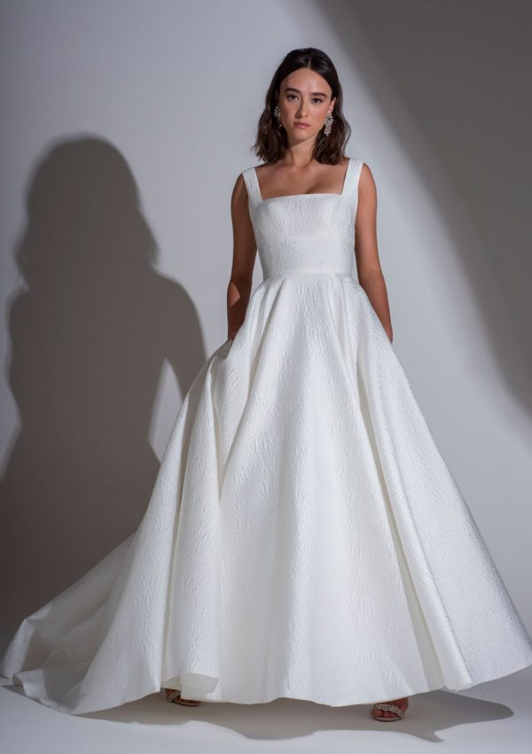 A woman stands against a plain background wearing the Freda Bennet Milan Wedding Dress from Blessings Of Brighton Wedding Dress Shop, featuring a square neckline and an A-line skirt with pockets. Her hair is styled down, casting a shadow on the wall behind her, as she looks directly at the camera with a serious expression.