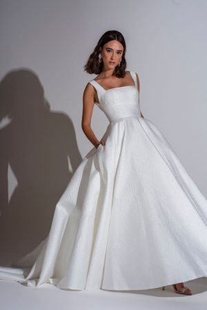 A woman stands against a plain background, wearing a sleeveless Freda Bennet - Milan Wedding Dress with a textured white gown, featuring an A-line skirt and square neckline. She has shoulder-length hair and poses with her hands in the pockets of her dress, casting a shadow on the wall behind her.