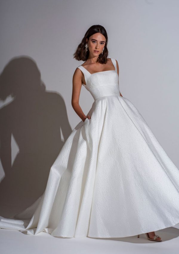 A woman stands against a plain background, wearing a sleeveless Freda Bennet - Milan Wedding Dress with a textured white gown, featuring an A-line skirt and square neckline. She has shoulder-length hair and poses with her hands in the pockets of her dress, casting a shadow on the wall behind her.