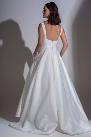 A woman stands with her back to the camera, showcasing the Freda Bennet - Milan wedding dress, featuring a deep square neckline and a full, textured A-line skirt. The dress has buttons running down the back, and she places her hands in its pockets. Her hair is styled in loose waves. Find it at Blessings Of Brighton Wedding Dress Shop.