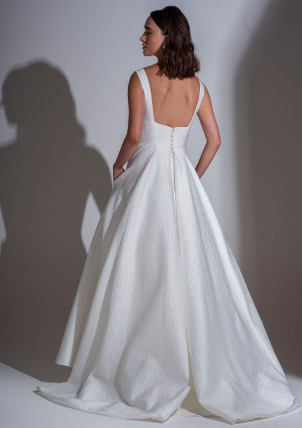 A woman stands with her back to the camera, showcasing the Freda Bennet - Milan wedding dress, featuring a deep square neckline and a full, textured A-line skirt. The dress has buttons running down the back, and she places her hands in its pockets. Her hair is styled in loose waves. Find it at Blessings Of Brighton Wedding Dress Shop.